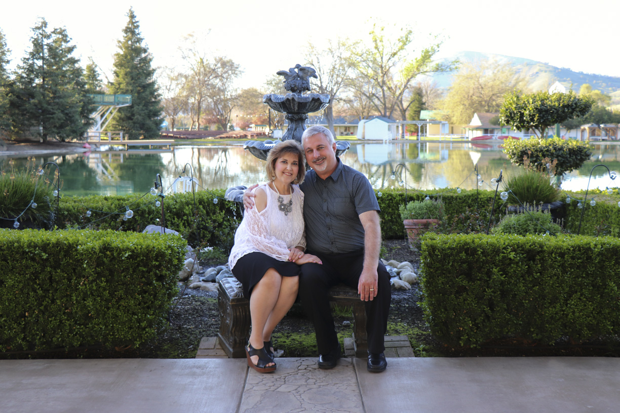 Paul and Rowena sitting together, posing for the camera with a fountain, water, and lush greenery in the background.