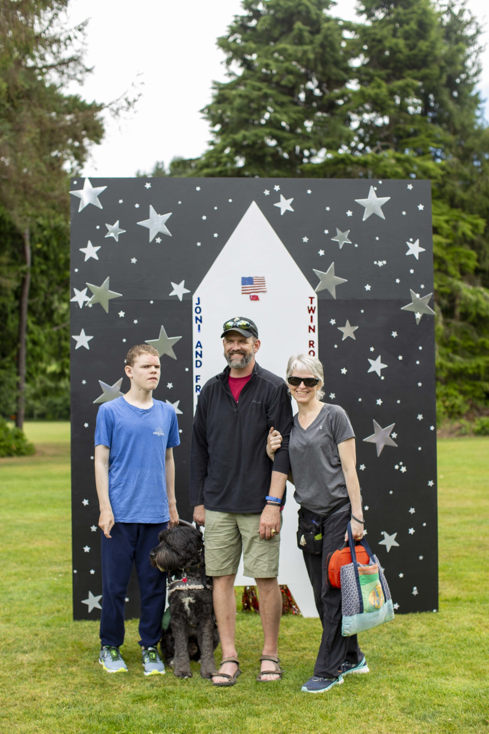 Noah, Will, JJ, and a dog smiling brightly and posing for the camera outdoors, with a rocket stand backdrop, enjoying their time at the Family Retreat.