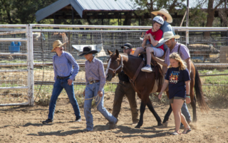 Brady sitting on a horse, looking happy and excited. Behind him, a male volunteer is assisting him, while five other volunteers walk alongside the horse, creating a supportive and joyful scene as they walk together.