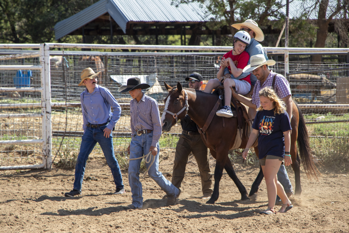 Brady sitting on a horse, looking happy and excited. Behind him, a male volunteer is assisting him, while five other volunteers walk alongside the horse, creating a supportive and joyful scene as they walk together.