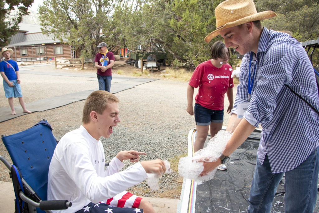 Brady sitting on a chair, joyfully playing with bubbles, with a volunteer in front of him also playing with bubbles. Both are smiling and having fun. In the background, several people are visible, contributing to the cheerful atmosphere.