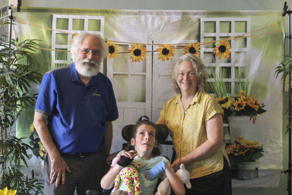 Mike and Mary are both standing next to Jessica, who is in her wheelchair. They are all smiling and posing for the camera at a Family Retreat, with a cute backdrop cloth featuring sunflowers and door and window panels creating a warm, inviting atmosphere.