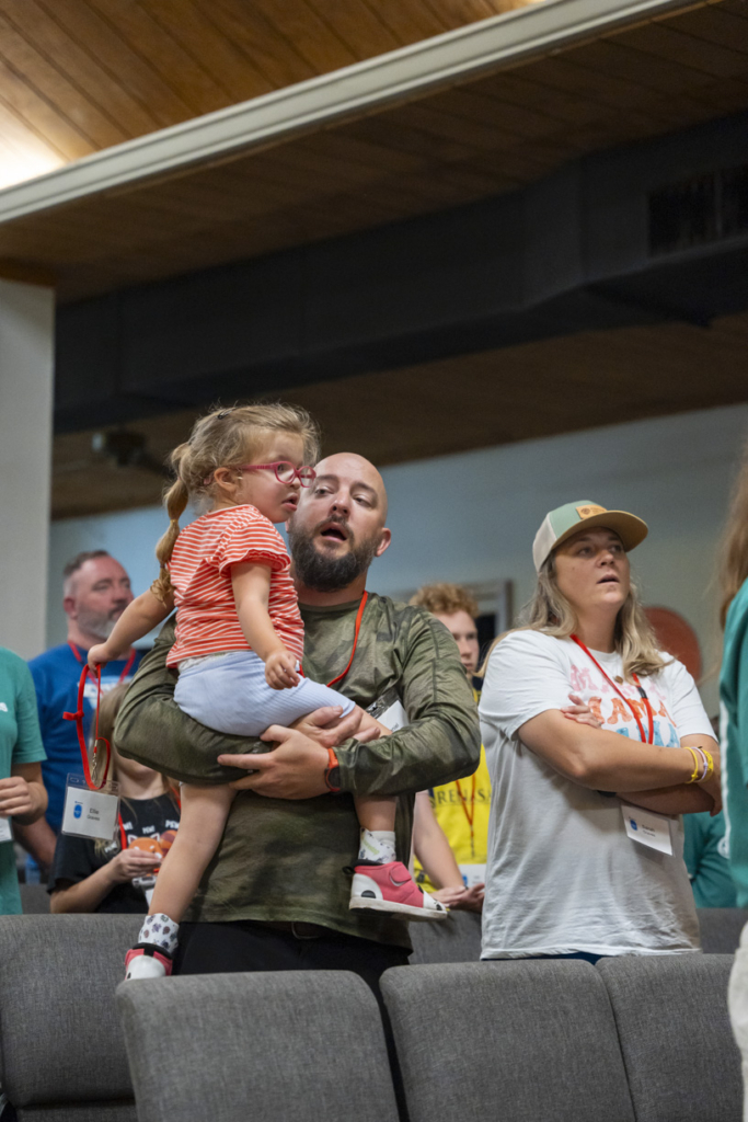 Daniel is looking at his daughter, Ellie, while carrying her in his arms as he stands next to his wife, Sarah. They are worshiping together with other attendees and volunteers at the Family Retreat, creating a moment of shared connection and devotion.