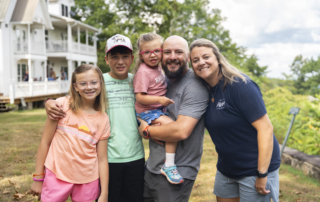 Abby, Sam, Ellie, Daniel, and Sarah pose and smile for the camera during the Family Retreat. Ellie is being carried by Daniel. Behind them is a house on the left, with trees adding to the peaceful outdoor setting.