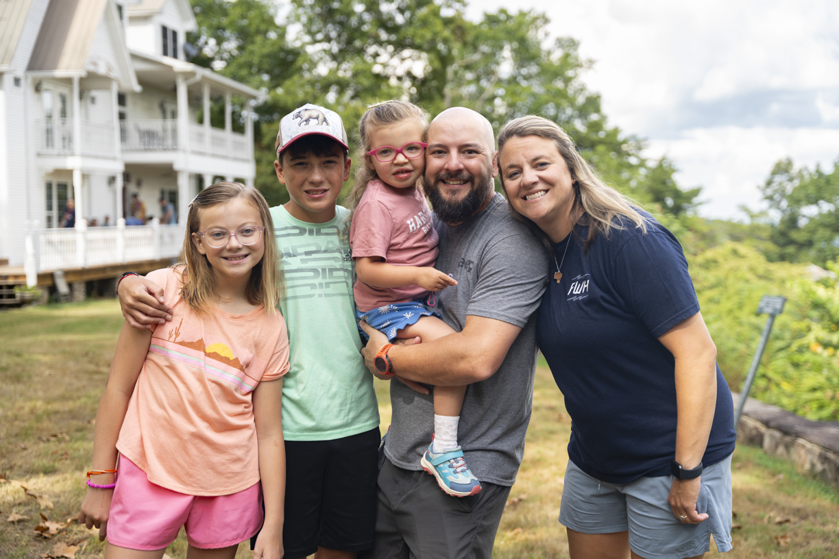 Abby, Sam, Ellie, Daniel, and Sarah pose and smile for the camera during the Family Retreat. Ellie is being carried by Daniel. Behind them is a house on the left, with trees adding to the peaceful outdoor setting.