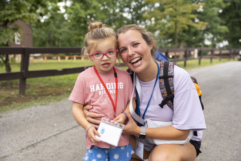 Ellie stands beside her buddy, Viktoriya, at a Family Retreat. Both are smiling happily and posing for the camera, radiating a joyful energy. In the background, there is green grass surrounded by lush trees, creating a peaceful outdoor atmosphere.