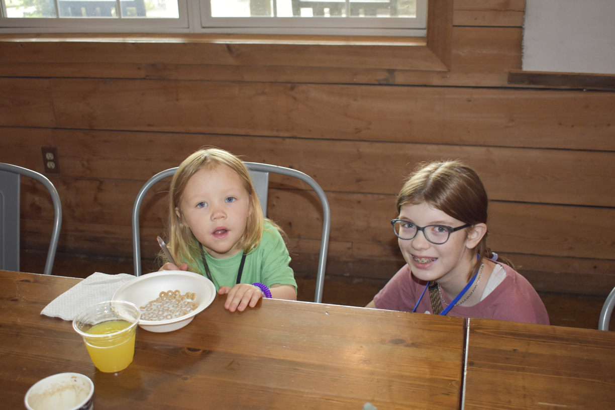 Alexa Anne is smiling and looking at the camera, with a little girl sitting beside her. On the table in front of them are a bowl of cereal and a glass of orange juice.