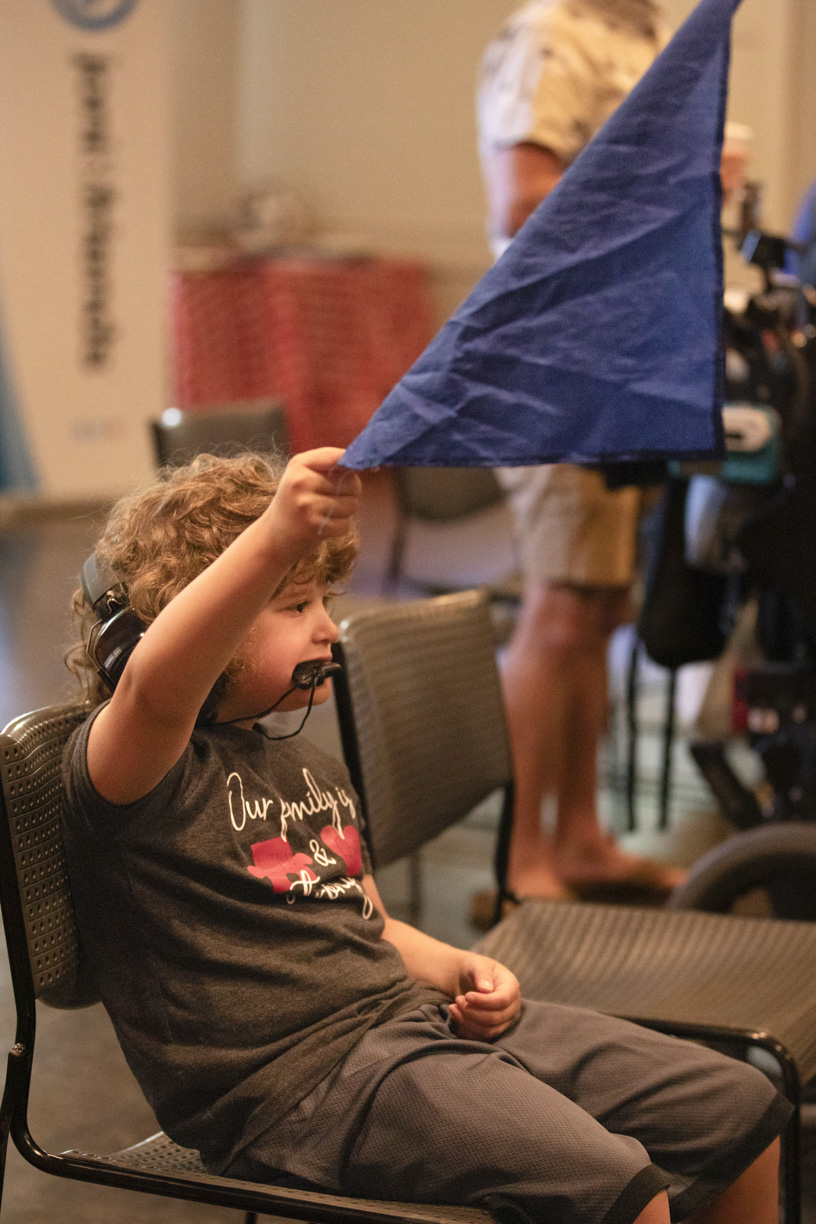 Elijah is sitting on a chair, holding a blue flag and raising it.