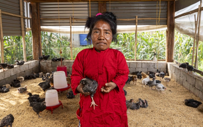 Kalyani standing and looking at the camera while holding a chicken inside her chicken coop, built by Joni and Friends, with chickens scattered around.