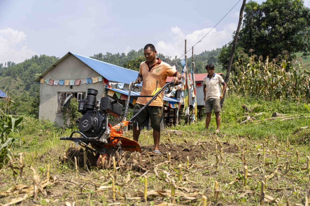 Biraj working the land with a new electric tractor preparing the soil for corn cultivation. A man stands nearby, observing the tilling process. In the background, there is a house and several trees, providing a peaceful rural setting.