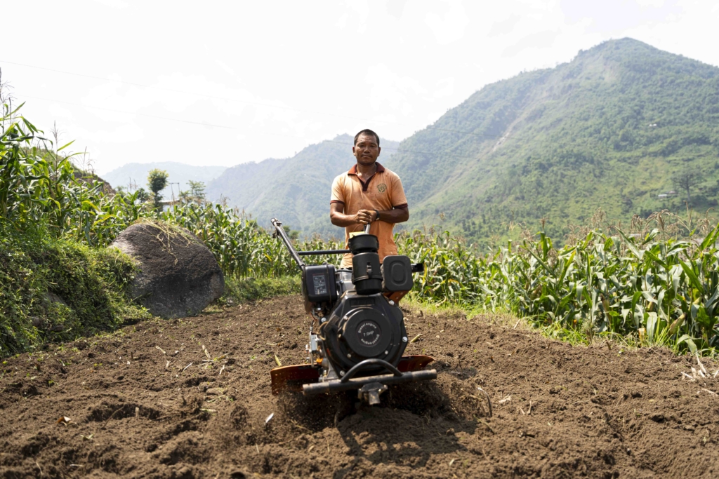 Biraj standing on the soil with his new electric tractor, surrounded by tall corn plants. In the background, the landscape is framed by majestic mountains, adding to the rural beauty of the scene.