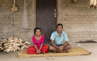 Padma and Biraj sitting next to each other on a straw mat, with ears of corn piled on their left side. Both of them are looking at the camera.