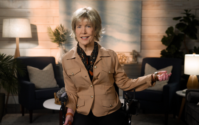 Joni Eareckson Tada smiling for the camera while wearing a brown safari jacket, a black scarf with floral details, in a cozy living room.