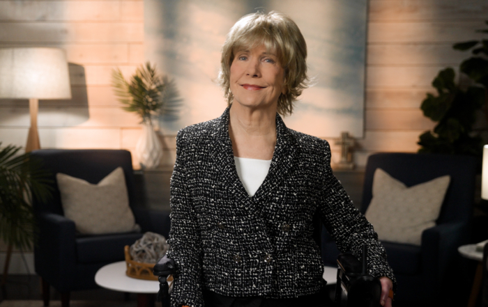 Joni Eareckson Tada smiles for the camera while sitting in her wheelchair, wearing a black blazer with white boucle over a white top, in a cozy living room setting.