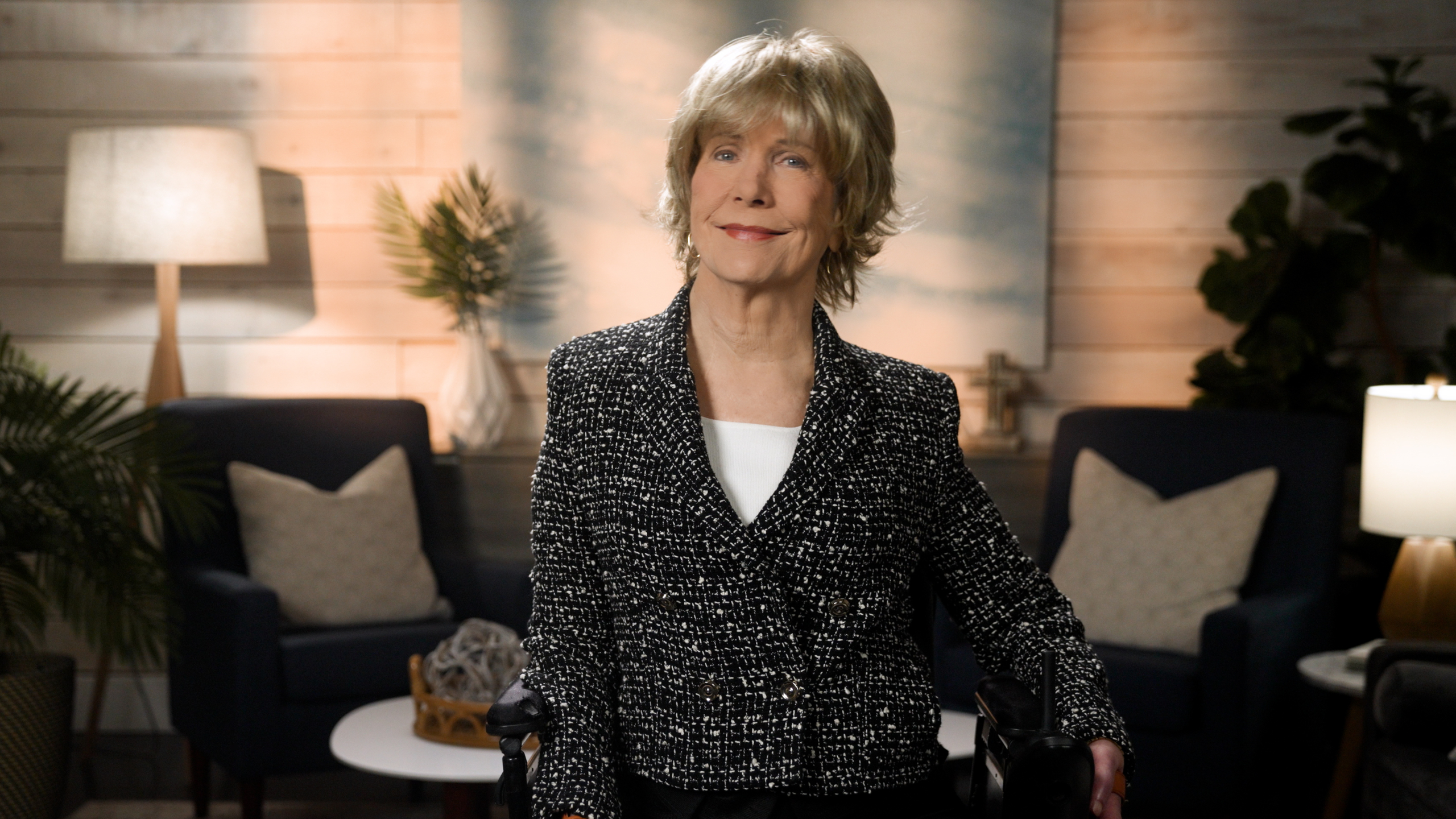 Joni Eareckson Tada smiles for the camera while sitting in her wheelchair, wearing a black blazer with white boucle over a white top, in a cozy living room setting.