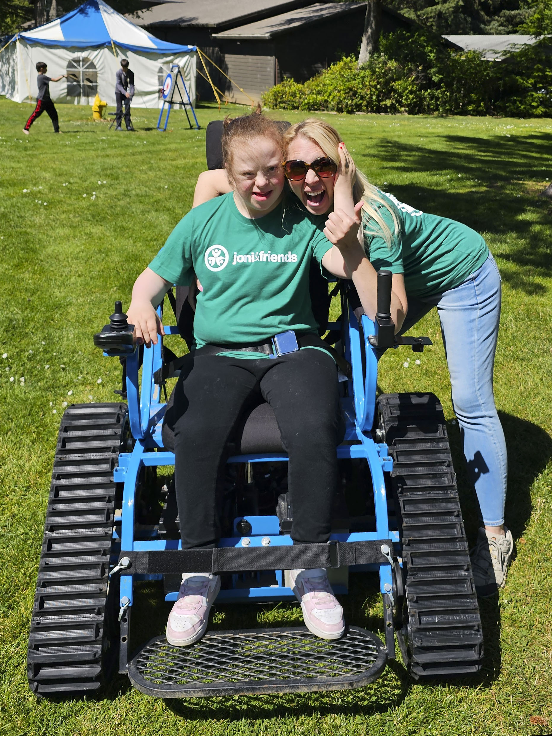 Sarah sitting in a rubber-tracked wheelchair while her mom, Shauna, hugs her. Both look happy during Family Retreat.