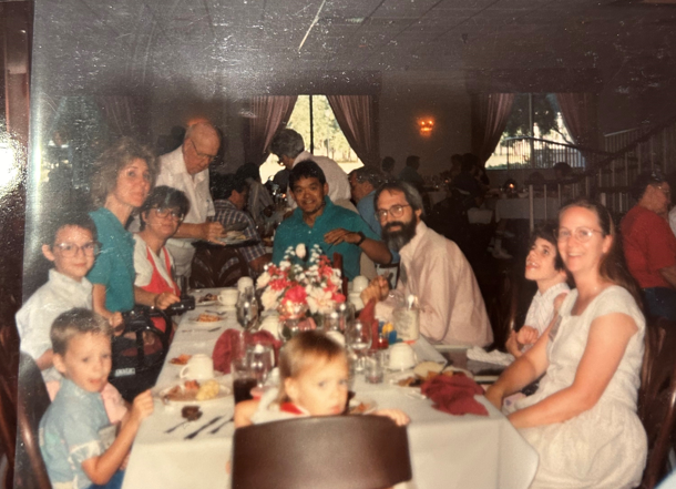 A historical photo of Mike, Jessica, Mary, Ken, Joni, another girl next to Joni, and three kids sitting together at a long table, all smiling and posing for the camera. In the background, other people are enjoying their time, chatting and eating at a Family Retreat.