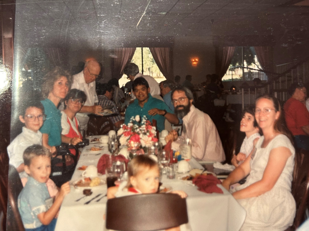 A historical photo of Mike, Jessica, Mary, Ken, Joni, another girl next to Joni, and three kids sitting together at a long table, all smiling and posing for the camera. In the background, other people are enjoying their time, chatting and eating at a Family Retreat.