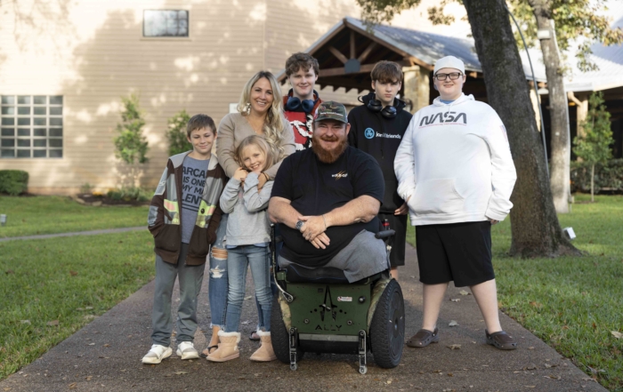 Jay sitting in his wheelchair surrounded by Anne and their five children (four boys and one girl) in an outdoor setting. Everyone looks happy.