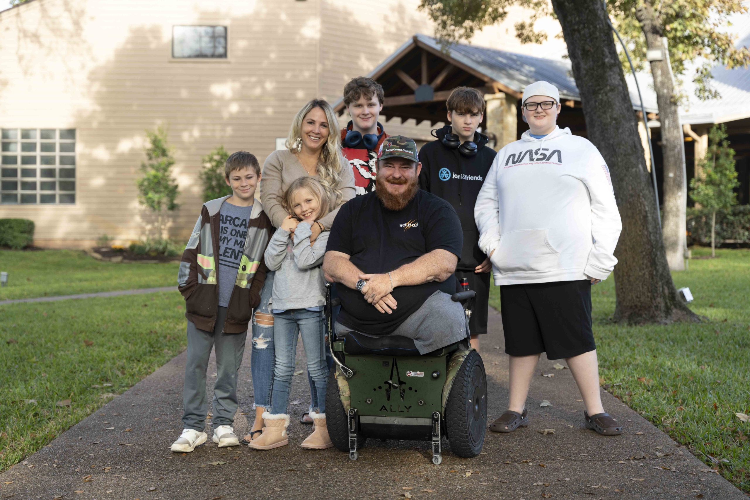 Jay sitting in his wheelchair surrounded by Anne and their five children (four boys and one girl) in an outdoor setting. Everyone looks happy.