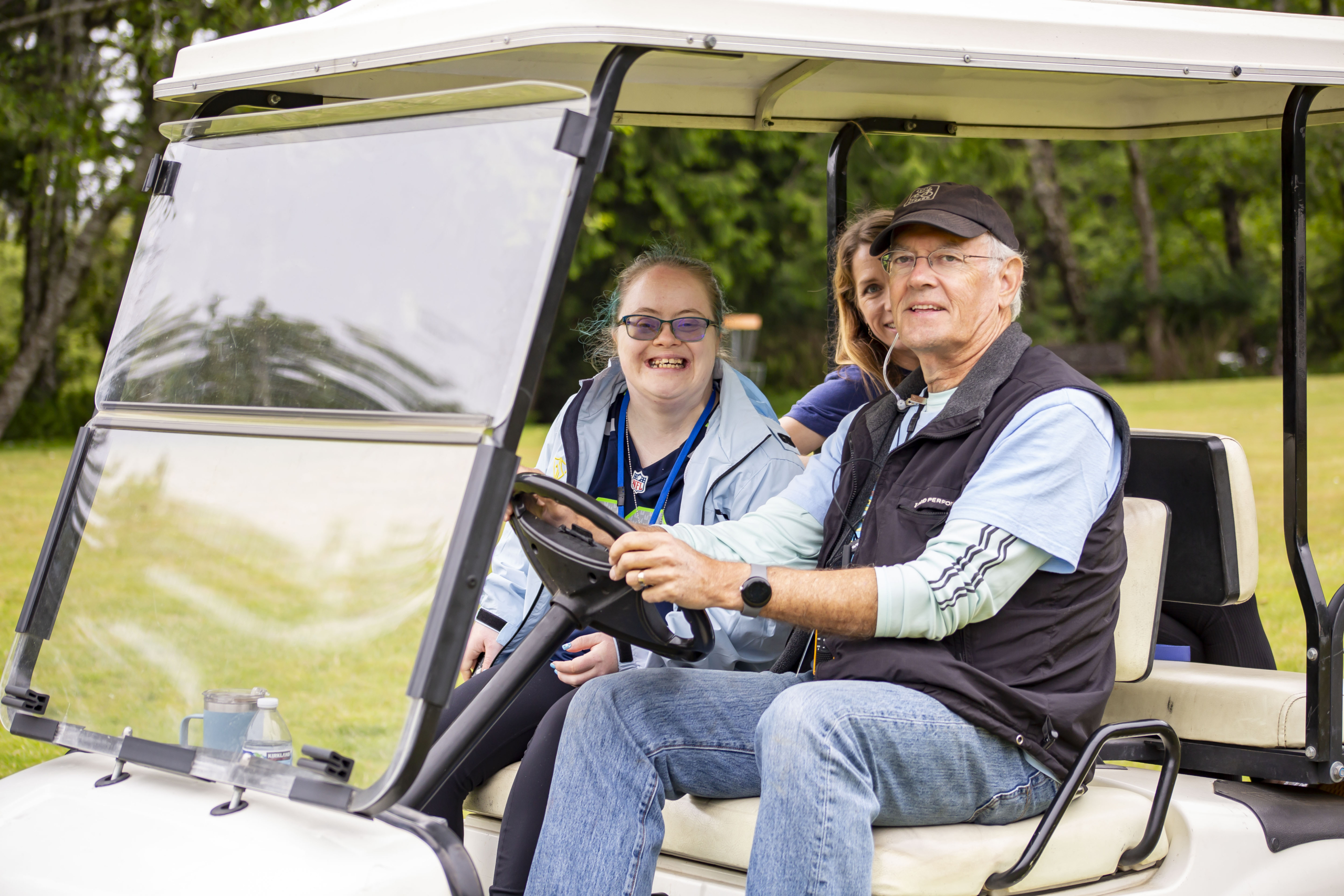 Emily Satterberg smiling while riding in a golf cart next to a man driving, with a woman sitting behind them.