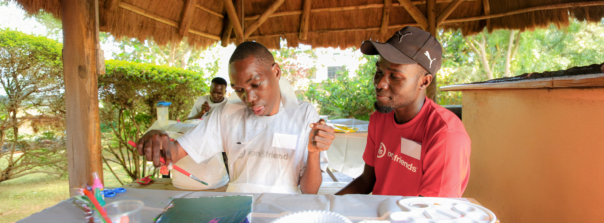 A young man paints with focus under a thatched pavilion, supported by a mentor in a red 'Joni and Friends' shirt, with lush greenery in the background.