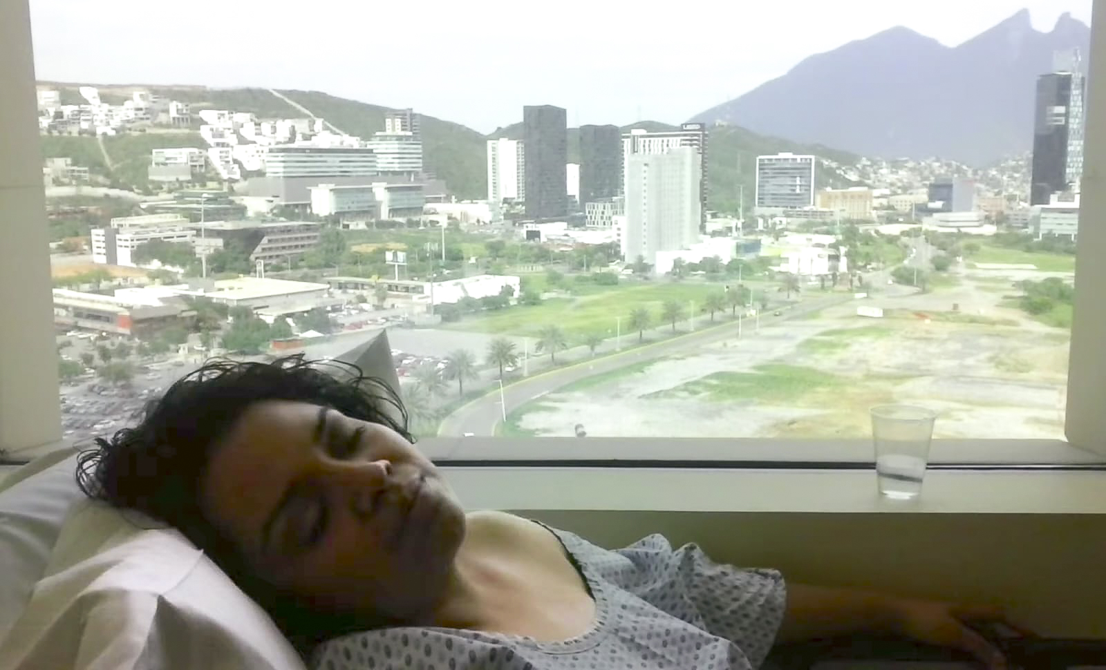 Vianey lying on a bed with her eyes closed, wearing a hospital gown and a beautiful outdoor view visible through her window.