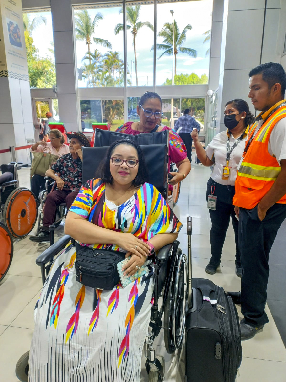 Vianey sitting in her wheelchair with a woman pushing her through a busy airport, with other travelers and airport staff visible in the background.