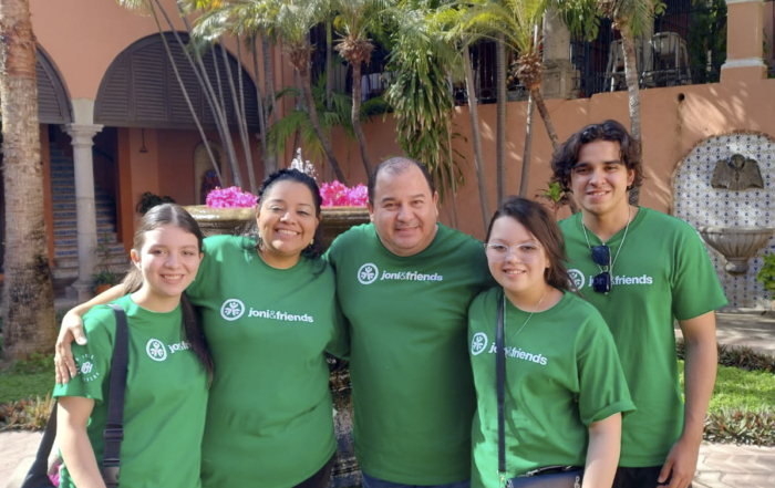 Vianey standing with her husband and three children (two girls and one boy) all wearing matching Joni and Friends t-shirts, with a scenic backdrop of beautiful coconut trees.
