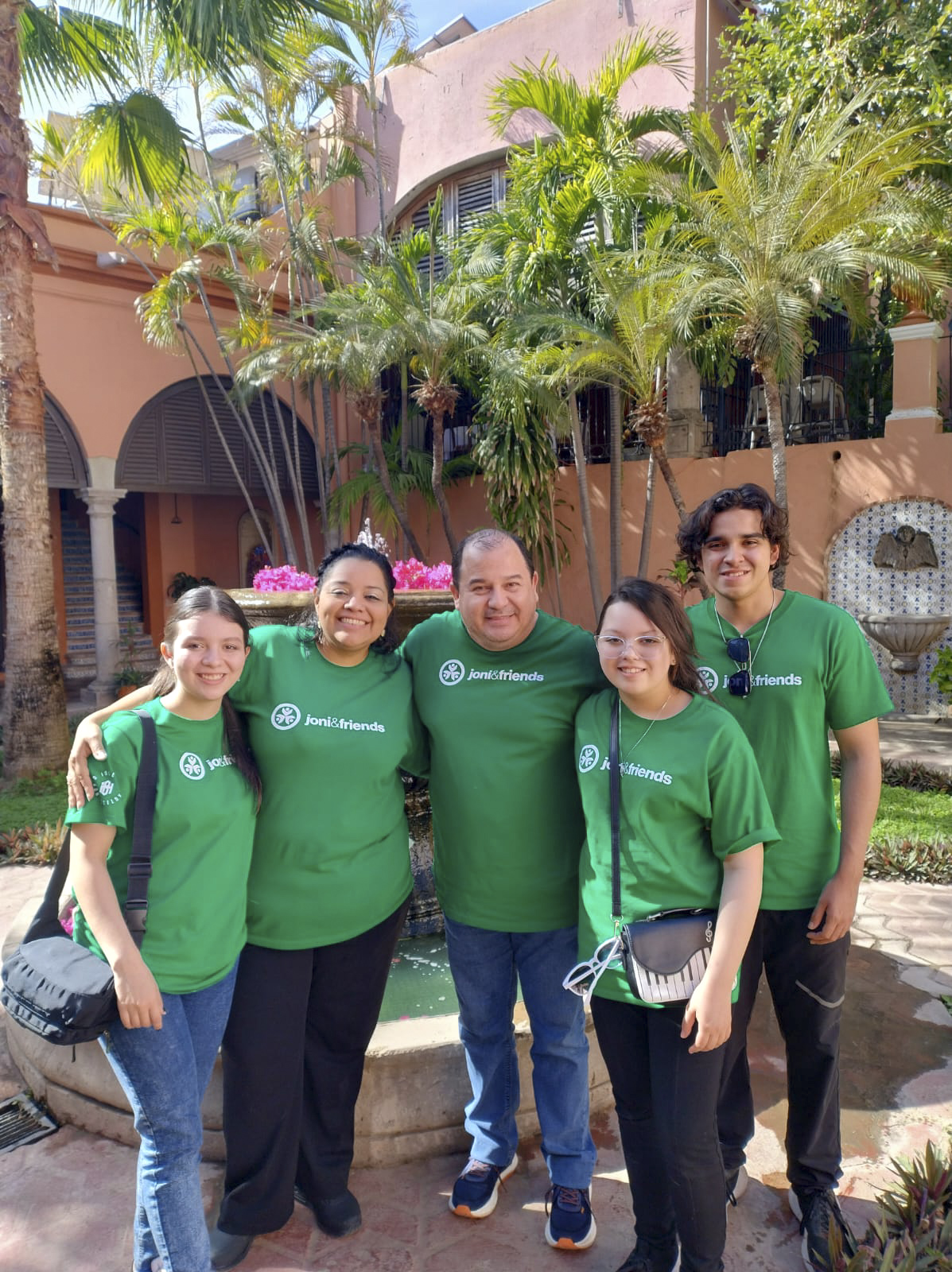 Vianey standing with her husband and three children (two girls and one boy) all wearing matching Joni and Friends t-shirts, with a scenic backdrop of beautiful coconut trees.