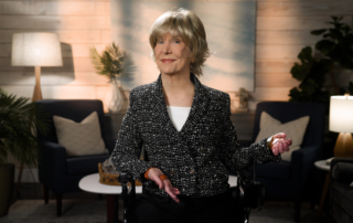 Joni Eareckson Tada smiles for the camera while sitting in her wheelchair, wearing a black blazer with white boucle over a white top, in a cozy living room setting.
