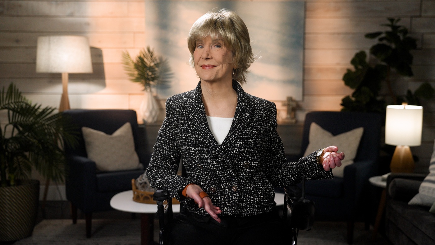 Joni Eareckson Tada smiles for the camera while sitting in her wheelchair, wearing a black blazer with white boucle over a white top, in a cozy living room setting.