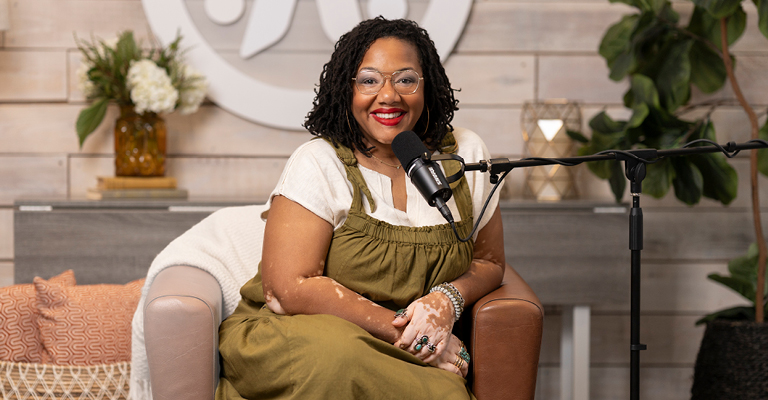 Stephanie Daniels sitting on a couch, smiling and posing for the camera with a microphone in front of her, in a cozy living room setup.