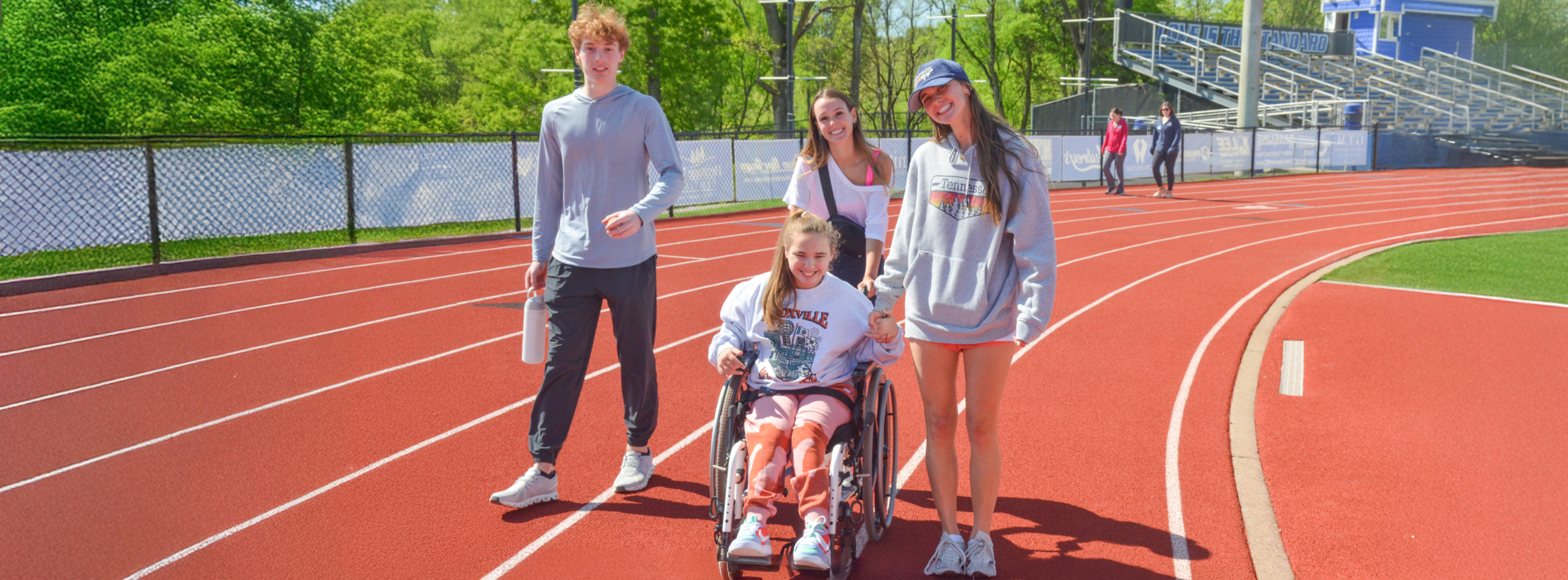 A group of four young people, three standing and one in a wheelchair, smile and walk together on a red running track surrounded by green trees on a sunny day.