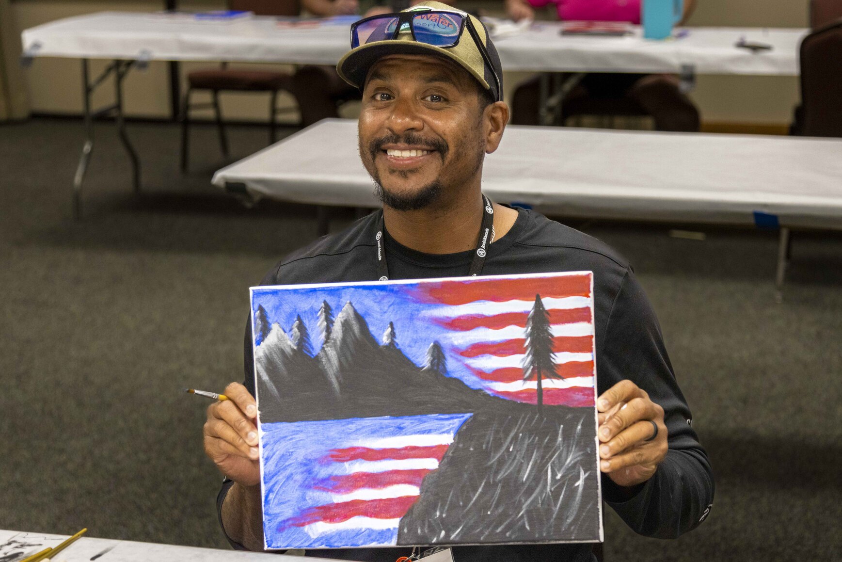 JB smiling for the camera while holding his artwork of an American flag with a silhouette of mountains and trees.