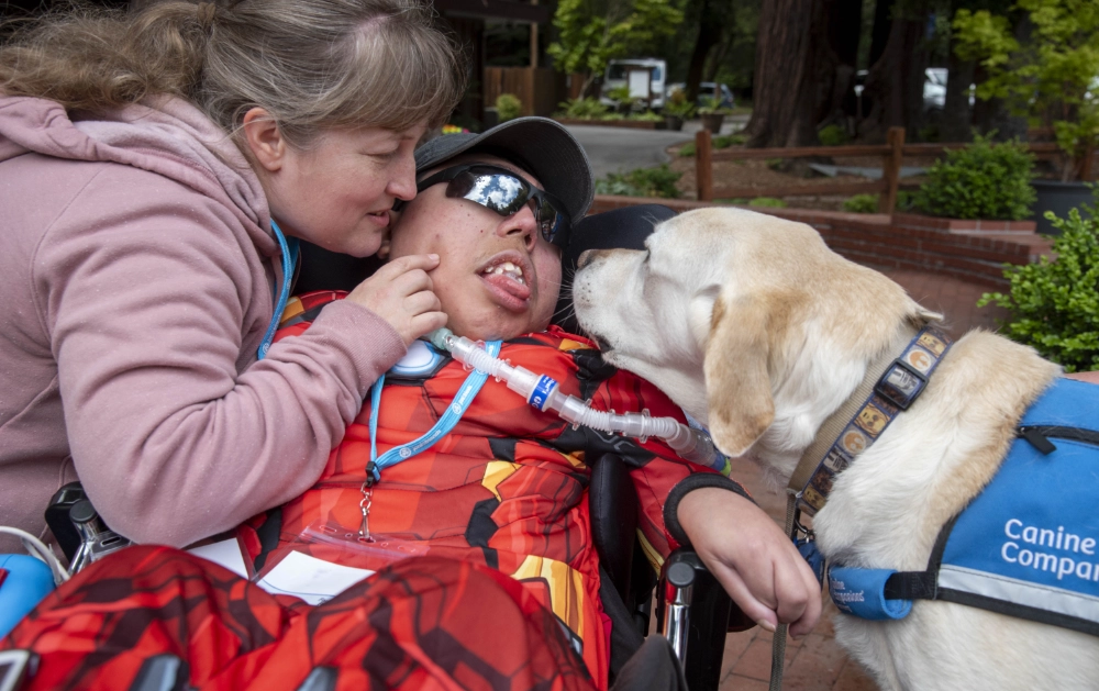 A woman is showing affection to a boy with sunglasses in a wheelchair as a service dog also joins