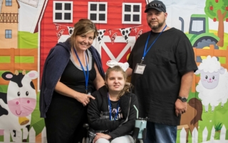 Tana sits in her wheelchair with her mom, Jennifer, and another man beside them. They smile and pose for the camera.