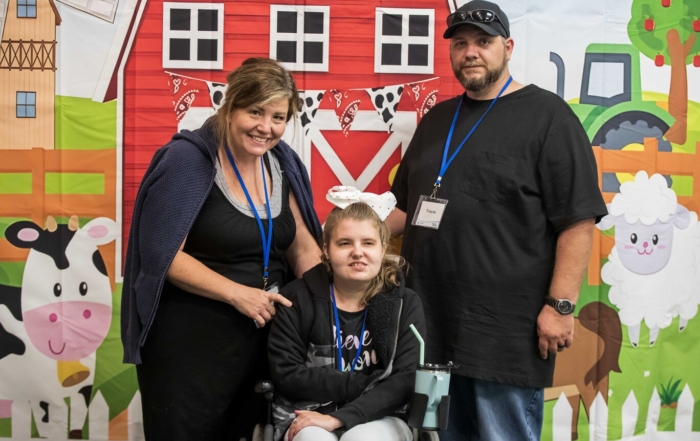 Tana sits in her wheelchair with her mom, Jennifer, and another man beside them. They smile and pose for the camera.