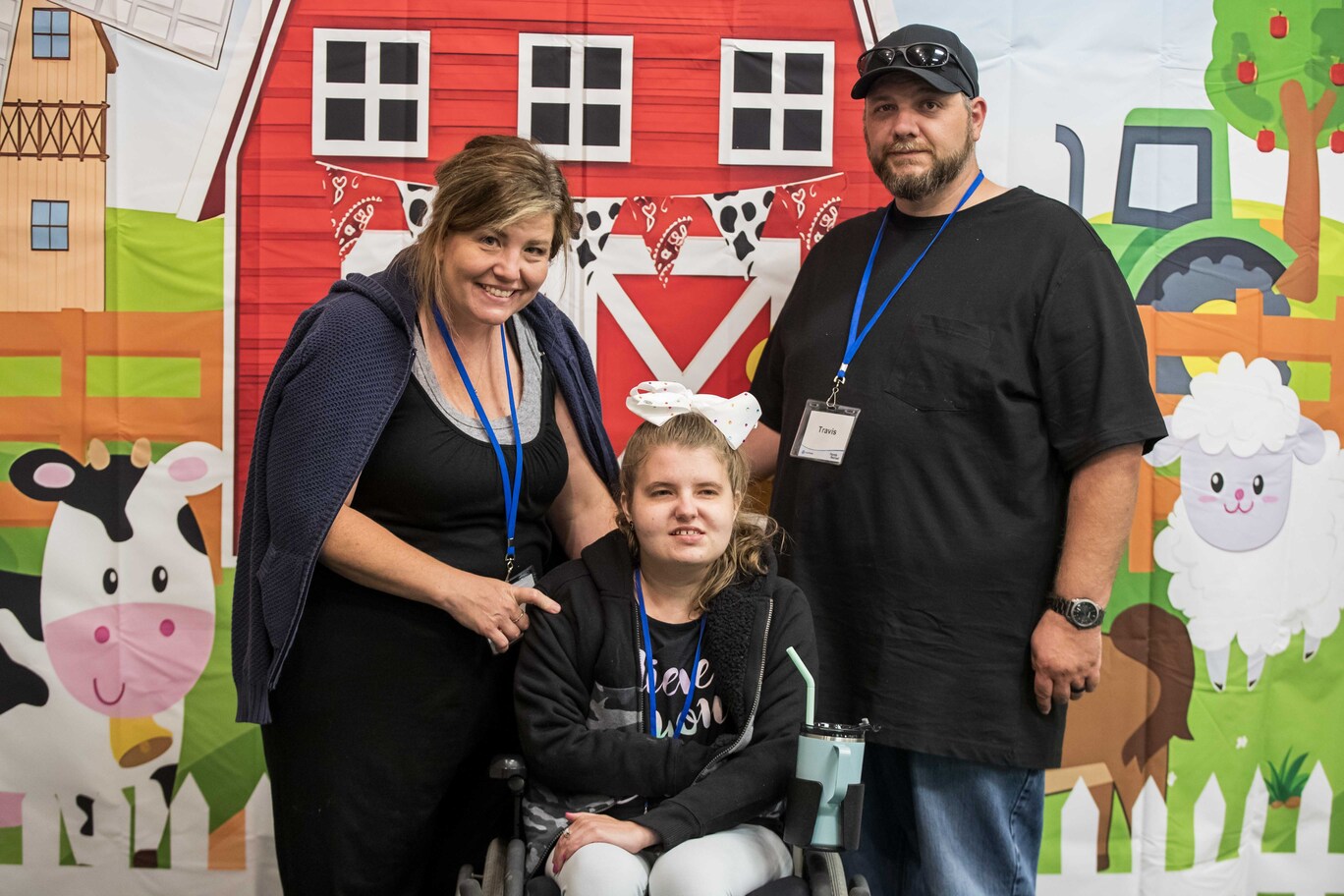 Tana sits in her wheelchair with her mom, Jennifer, and another man beside them. They smile and pose for the camera.