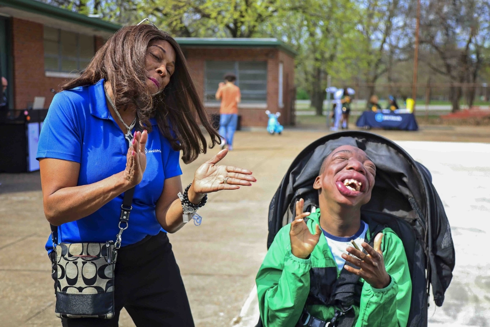 A woman in a blue shirt claps as a boy in his wheelchair claps along
