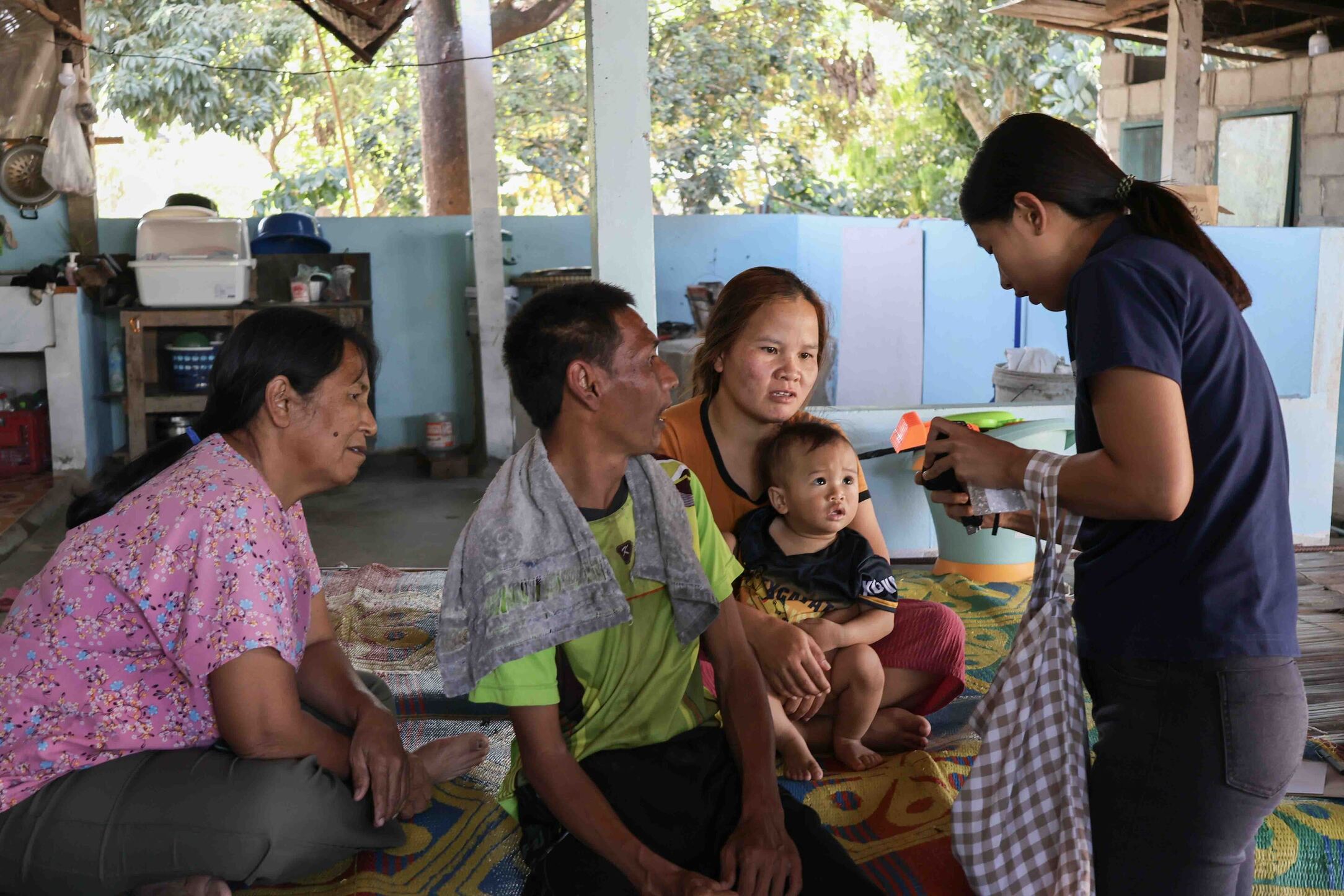 Janga sits on a bed next to her sister, who is holding her baby, and her sister's mother-in-law. A woman stands in front of them, showing them something, as they all look.