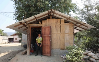 Janga standing by the door outside his home, smiling joyfully, with an overview of his home in the background.