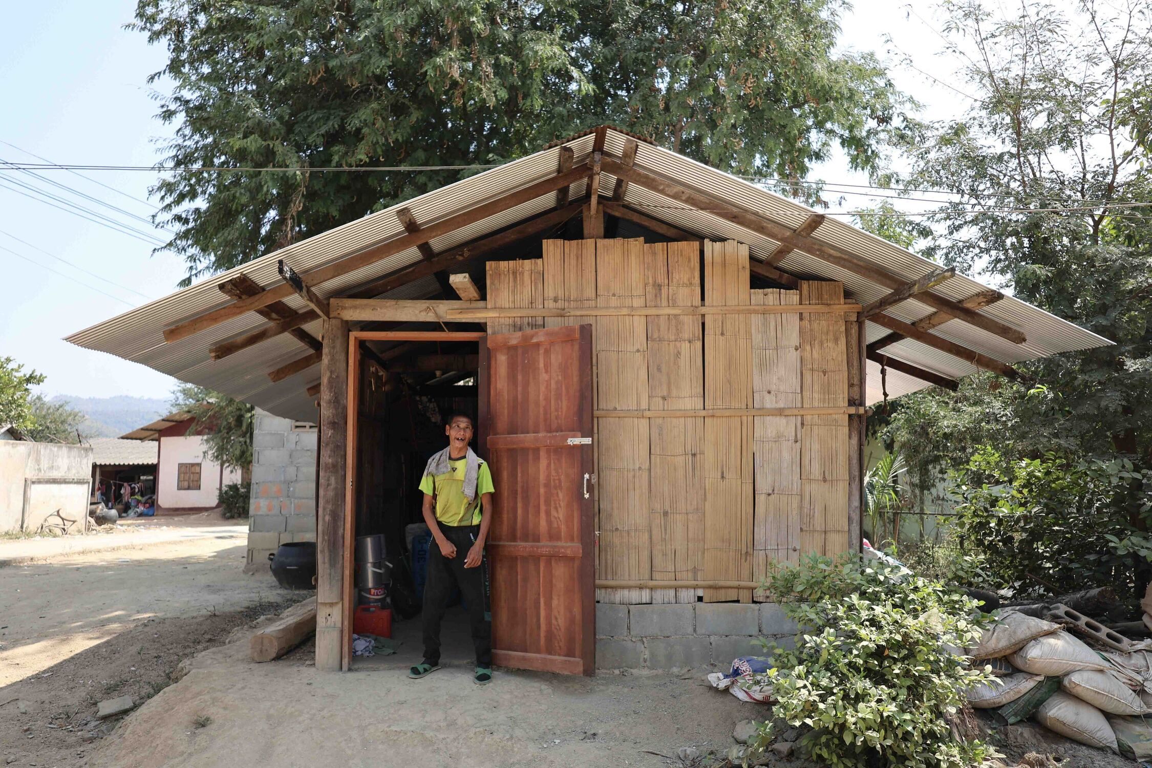 Janga standing by the door outside his home, smiling joyfully, with an overview of his home in the background.