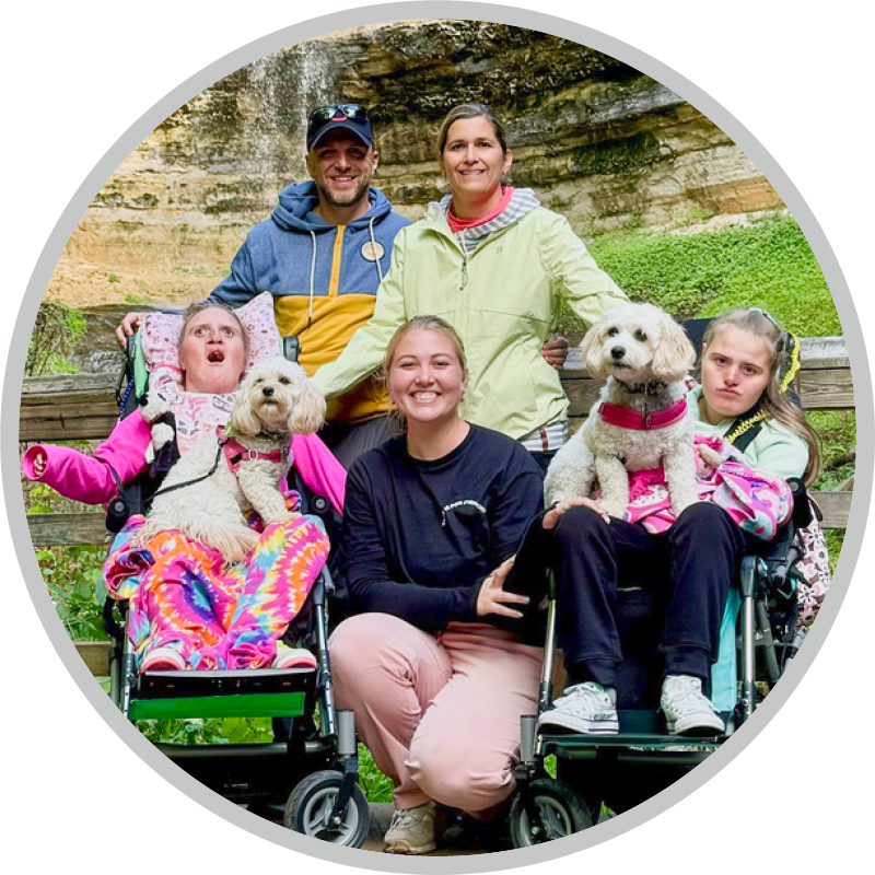 Paul and Kathy with another woman, along with their daughters Katelyn and Madelyn, each holding a dog on their laps, with a waterfall behind them.