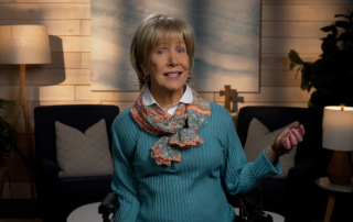 Joni Eareckson Tada smiling, wearing a long-sleeve shirt and a festive scarf in a cozy living room.