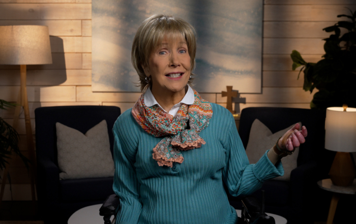 Joni Eareckson Tada smiling, wearing a long-sleeve shirt and a festive scarf in a cozy living room.