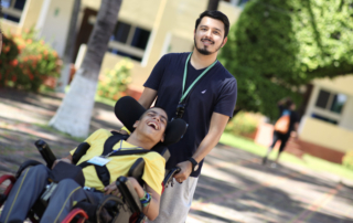 Jonathan Menjivar pushing a boy in his wheelchair, both smiling as they look at the camera.