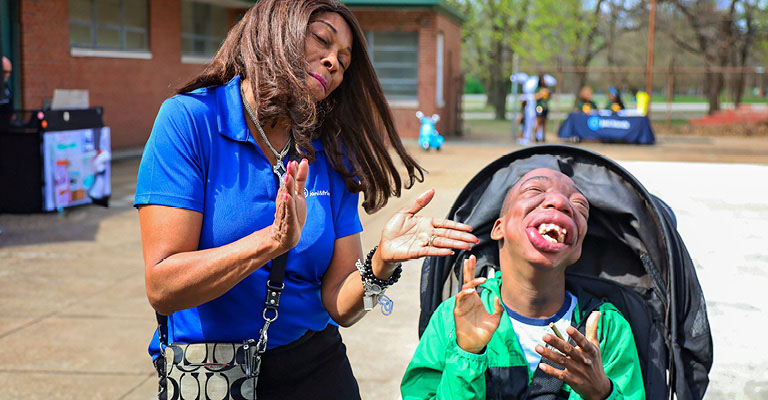 A staff member at Joni and Friends standing next to a boy in his stroller. Both are smiling joyfully and are clapping their hands.