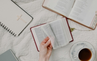 A close-up of a hand flipping through a Bible, with another Bible that is opened, a notebook, and a cup of coffee on the table.