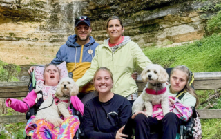 Paul and Kathy with another woman, along with their daughters Katelyn and Madelyn, each holding a dog on their laps, with a waterfall behind them.